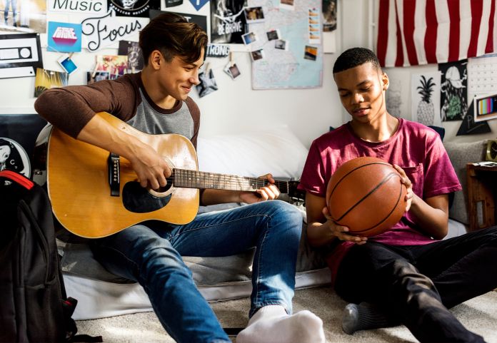 Teenage boys hanging out in a bedroom one holding a guitar and the other holding a basketball 