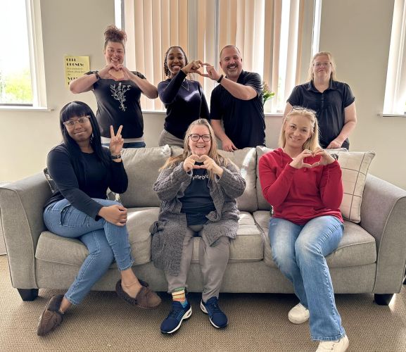 Team of employees sitting on couch and standing up holding hearts and peace signs